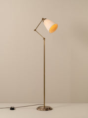 A mid-century modern floor lamp with a brass-colored adjustable metal stand, a pleated white shade with a warm yellow-gold interior, and a round stone base, shown against a neutral beige background.