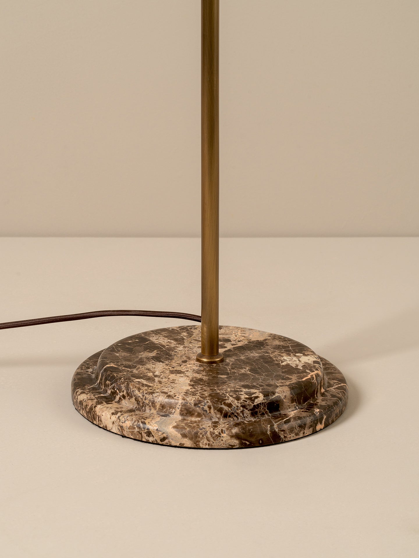 A close-up of a table lamp base featuring a round brown and beige marbled stone plate with a vertical brass-finished metal pole rising from the center, with a dark power cord extending to the left against a neutral beige background.