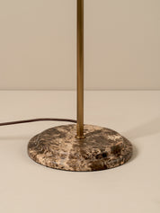 A close-up of a table lamp base featuring a round brown and beige marbled stone plate with a vertical brass-finished metal pole rising from the center, with a dark power cord extending to the left against a neutral beige background.
