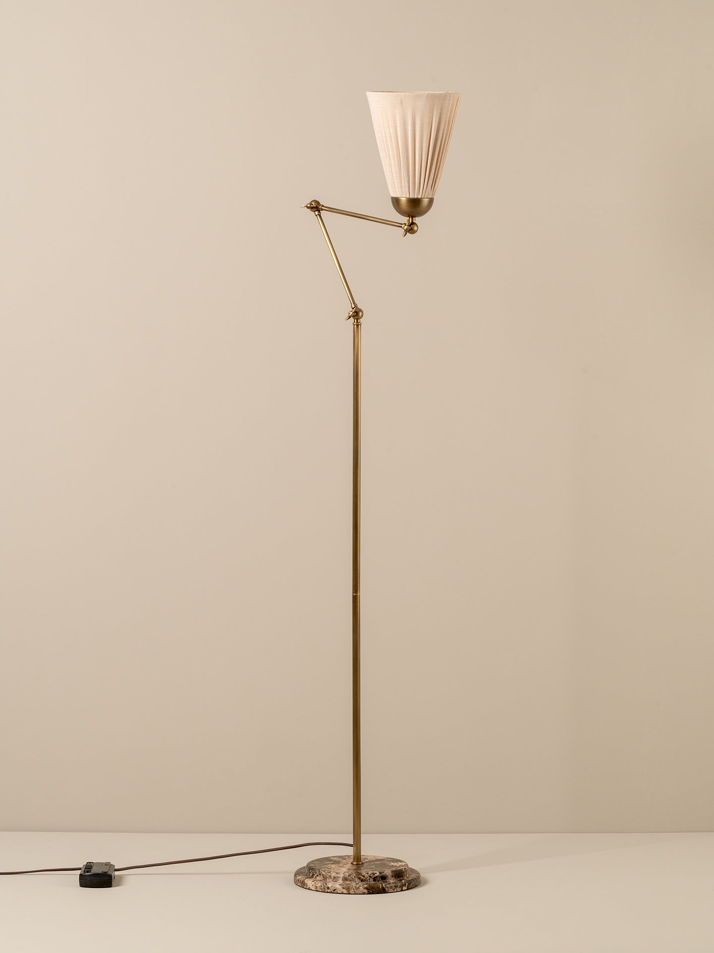 Mid-century modern floor lamp with a brass-colored metal frame, pleated off-white fabric shade, and a round patterned stone base, shown against a neutral beige background.