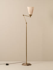 Mid-century modern floor lamp with a brass-colored metal frame, pleated off-white fabric shade, and a round patterned stone base, shown against a neutral beige background.