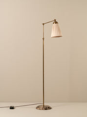 A mid-century modern floor lamp with a brass-colored metal stand, pleated off-white shade, and a round stone base, set against a neutral beige background.
