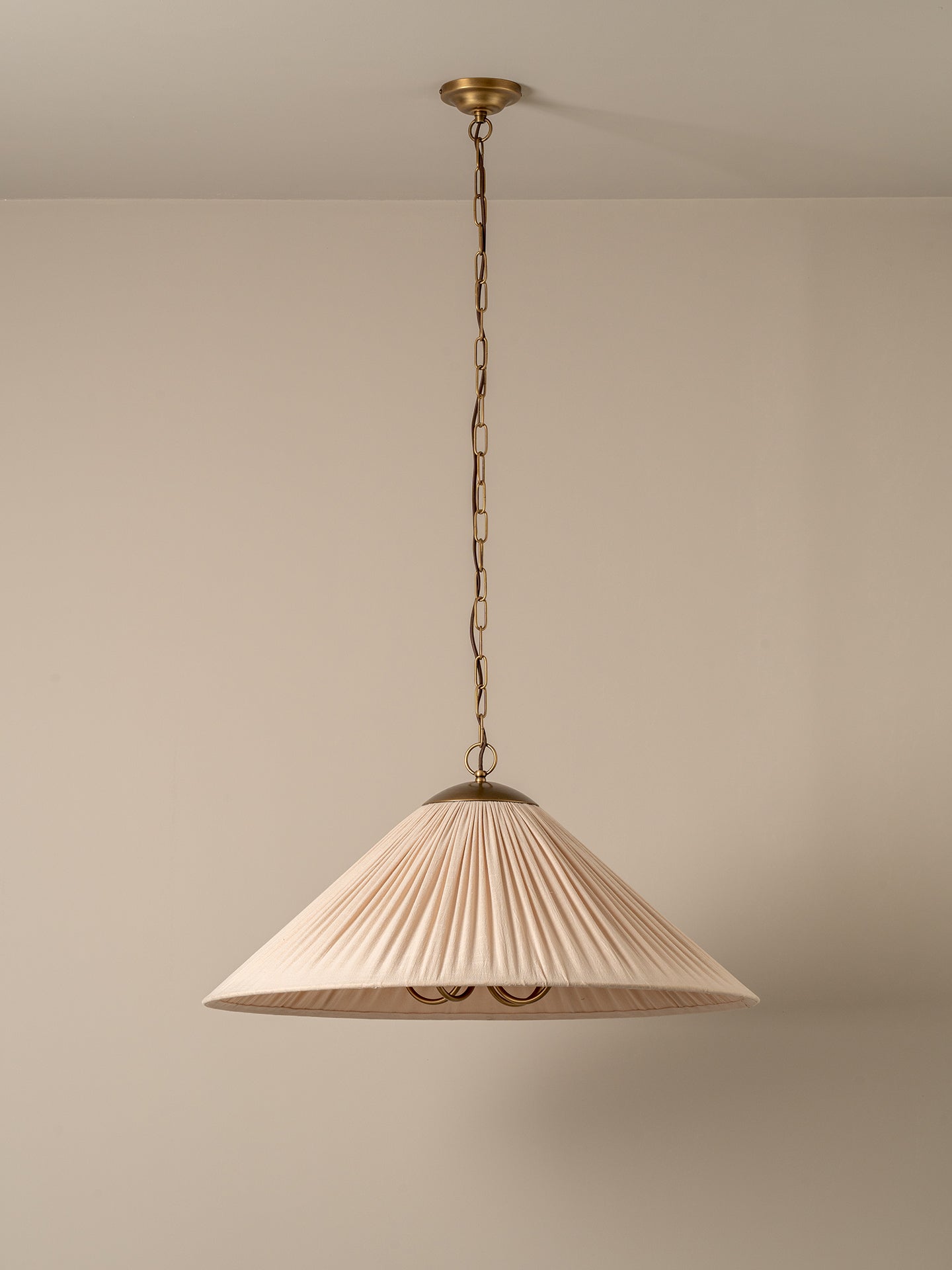 A pleated fabric pendant light shade in a warm cream or beige color, suspended from a brass chain and ceiling canopy against a neutral beige wall.