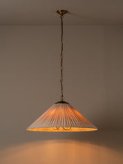 A hanging pendant light with a pleated white shade and warm brass-toned metal hardware, suspended against a plain wall. The light is turned on, casting a warm orange glow downward.