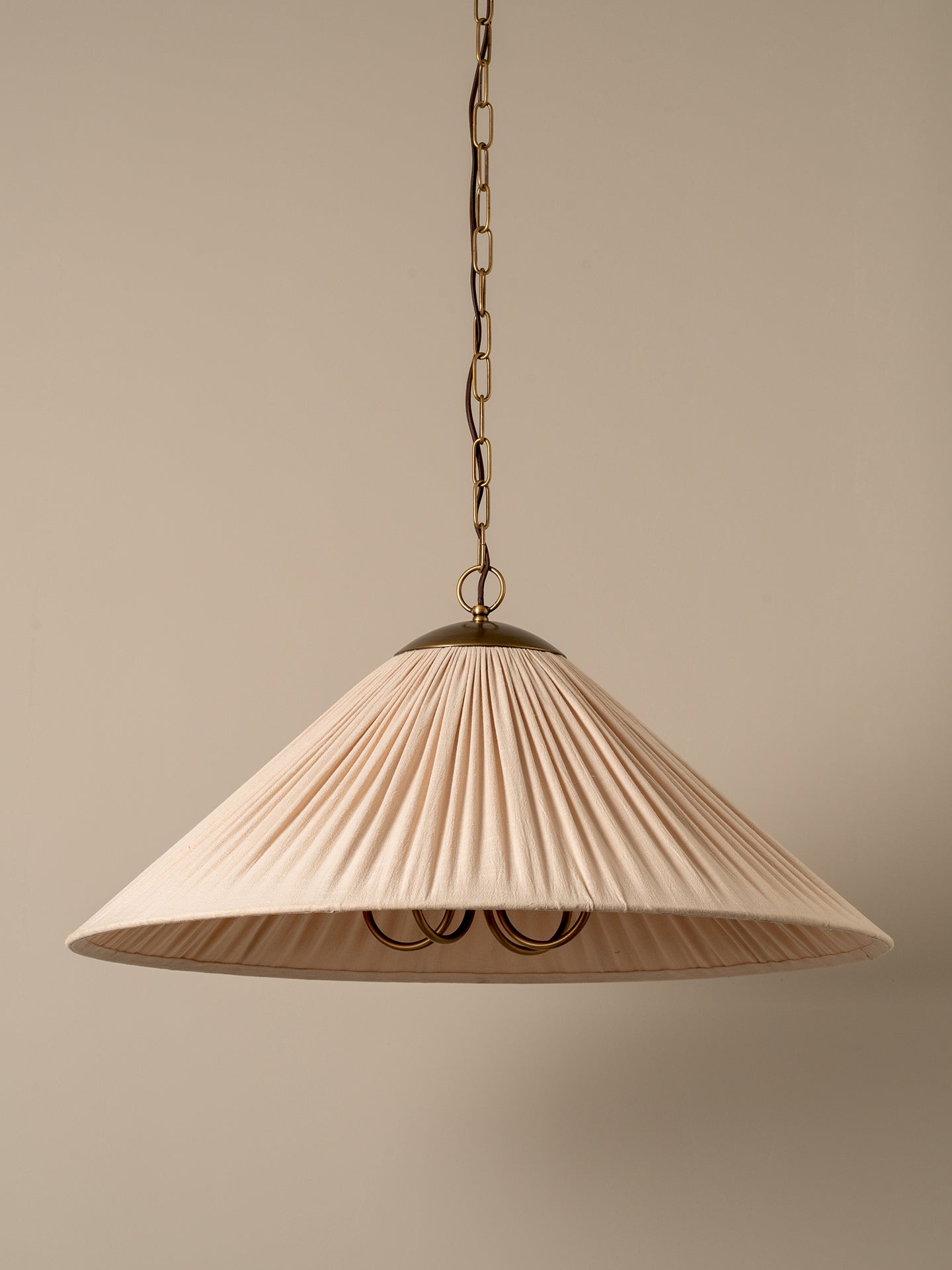 A pleated fabric pendant light shade in a warm beige or cream color, suspended from a brass chain against a solid beige background.