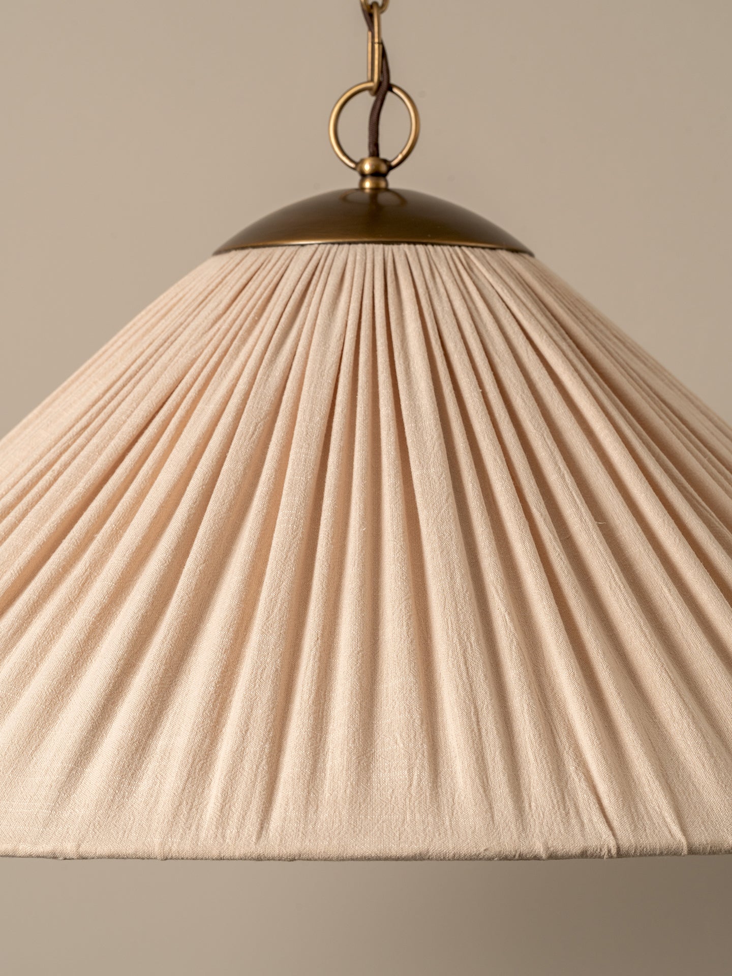 A close-up of a pleated fabric lampshade in a warm cream or beige color, suspended from a brass-colored metal ceiling mount against a neutral beige background.