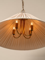 A modern chandelier pendant light with a large pleated fabric shade in a warm beige or blush tone, hanging from a brass-toned chain. The light has five candle-style bulbs with warm glowing filaments mounted on curved brass arms.