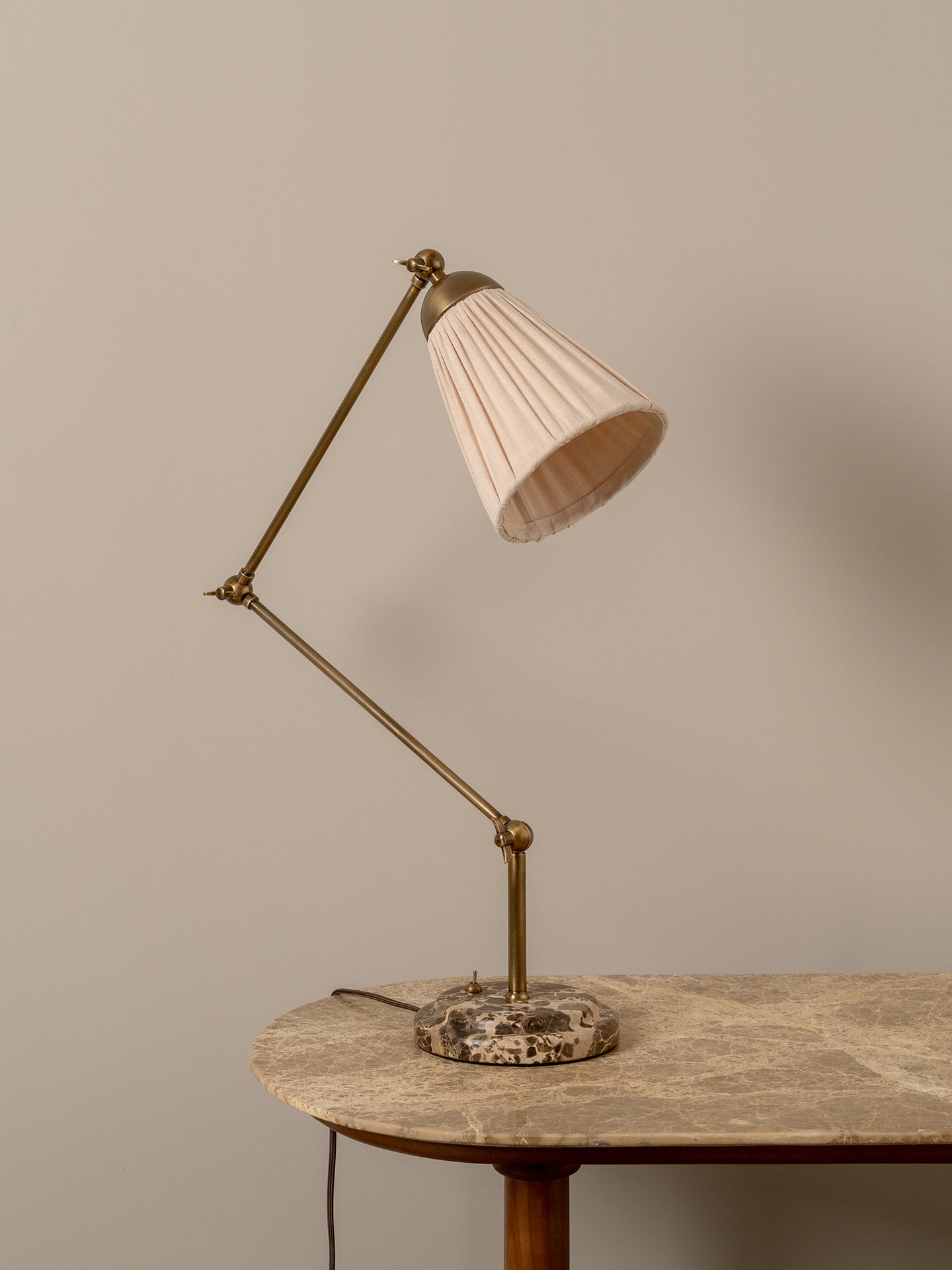 A mid-century modern adjustable desk lamp with a pleated cream shade, brass-colored metal arm, and a round stone base, shown on a stone-topped table against a neutral beige wall.