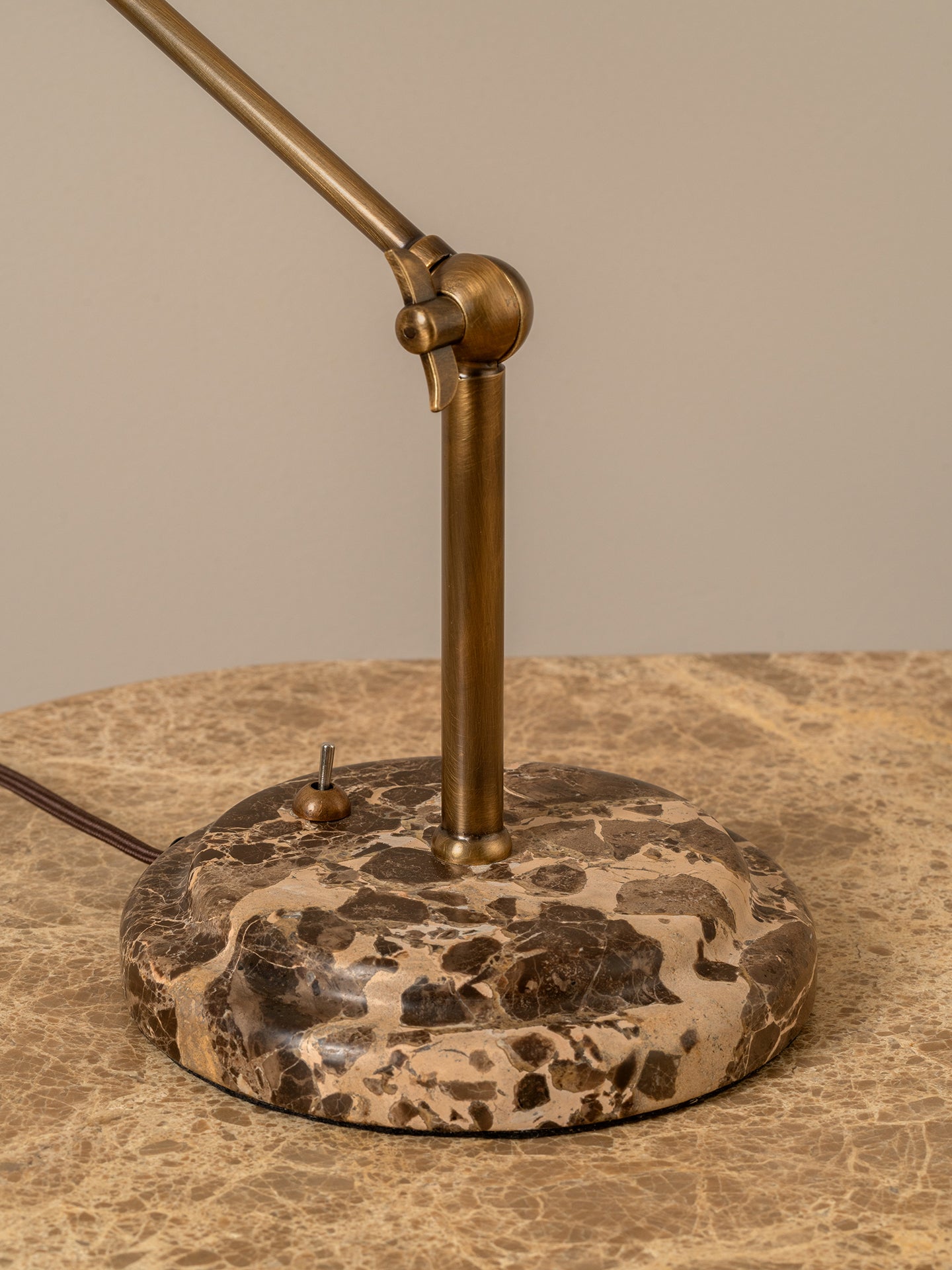 A close-up of a table lamp with a round dark brown and beige marbled base and a brass-colored metal arm, resting on a matching marble surface against a neutral beige background.
