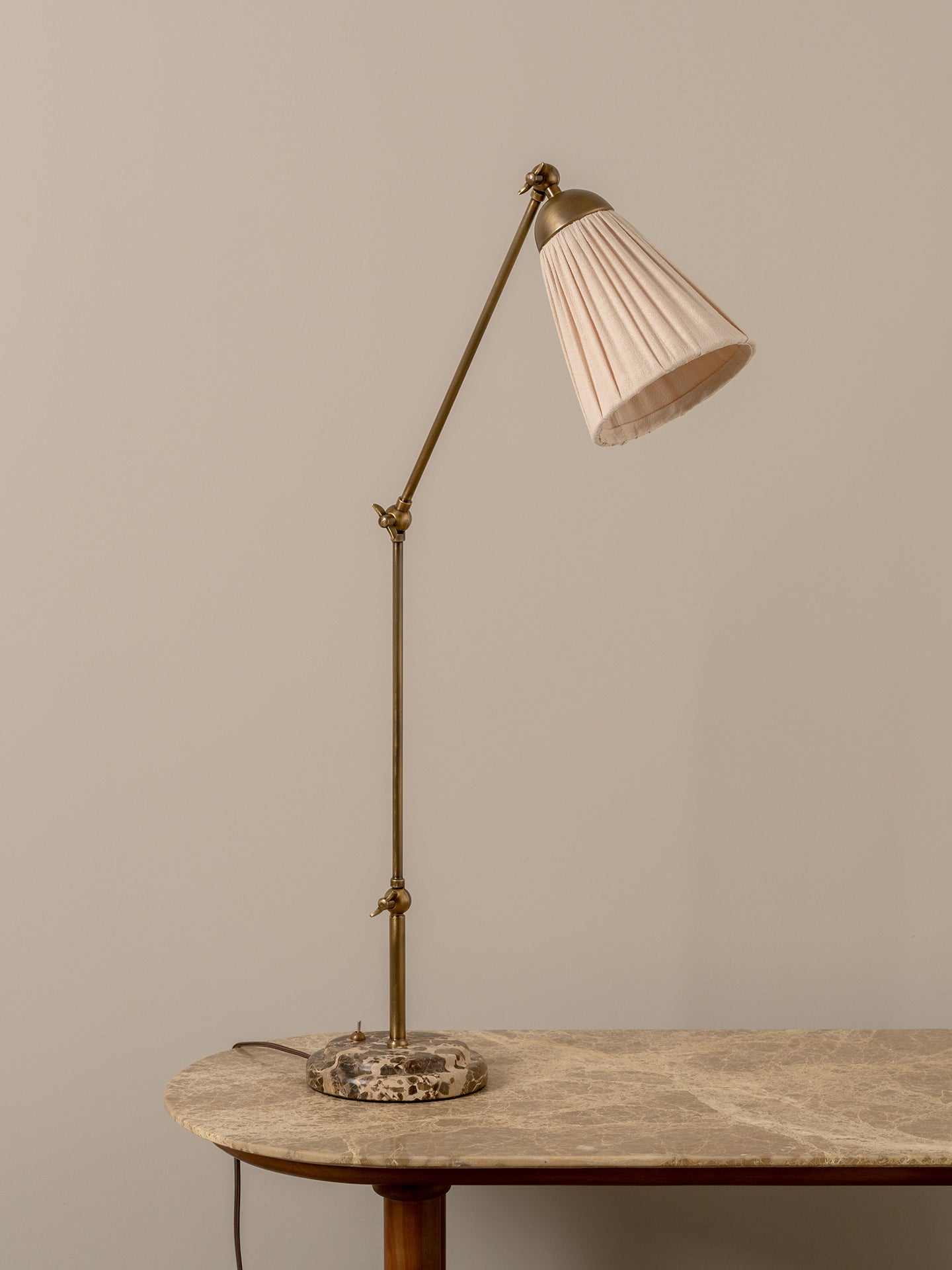 A mid-century modern adjustable desk lamp with a brass-colored metal stand and a pleated off-white shade, resting on a marble-topped table against a neutral beige wall.