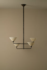 A modern multi-light chandelier with a dark metal frame and four cream pleated conical shades, suspended from a ceiling against a warm beige wall.
