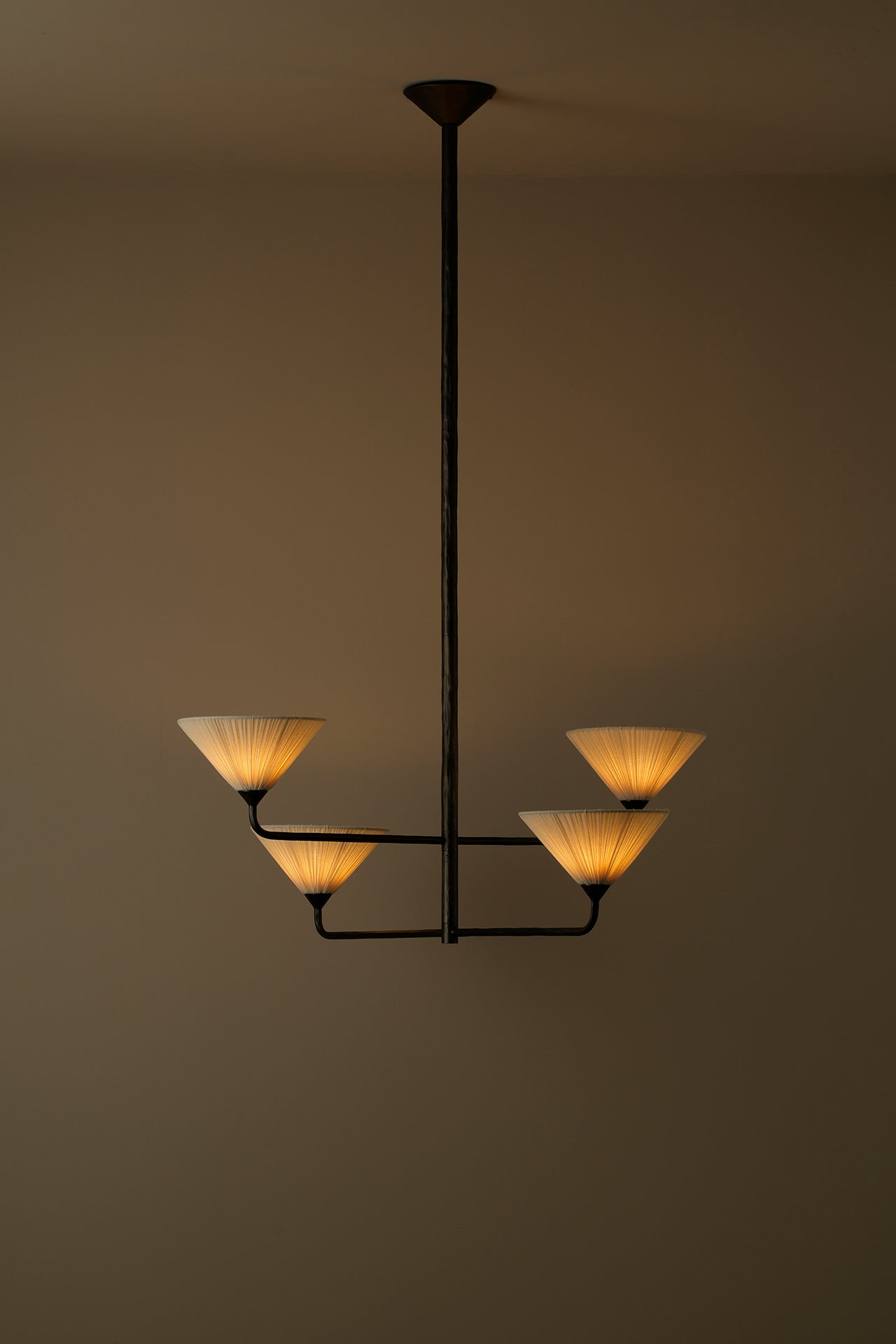 A modern four-light chandelier with a dark metal frame and warm glowing pleated shades, suspended against a plain warm brown wall.