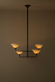 A modern four-light chandelier with a dark metal frame and warm glowing pleated shades, suspended against a plain warm brown wall.