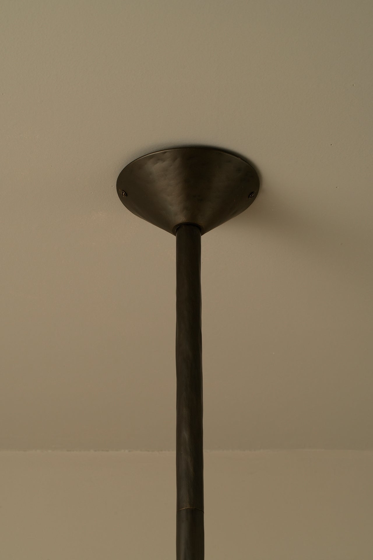 A close-up of a dark metal ceiling mount or pendant fixture ceiling canopy with a vertical rod extending downward against a plain beige wall.