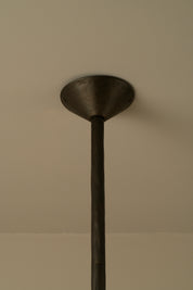 A close-up of a dark metal ceiling mount or pendant fixture ceiling canopy with a vertical rod extending downward against a plain beige wall.