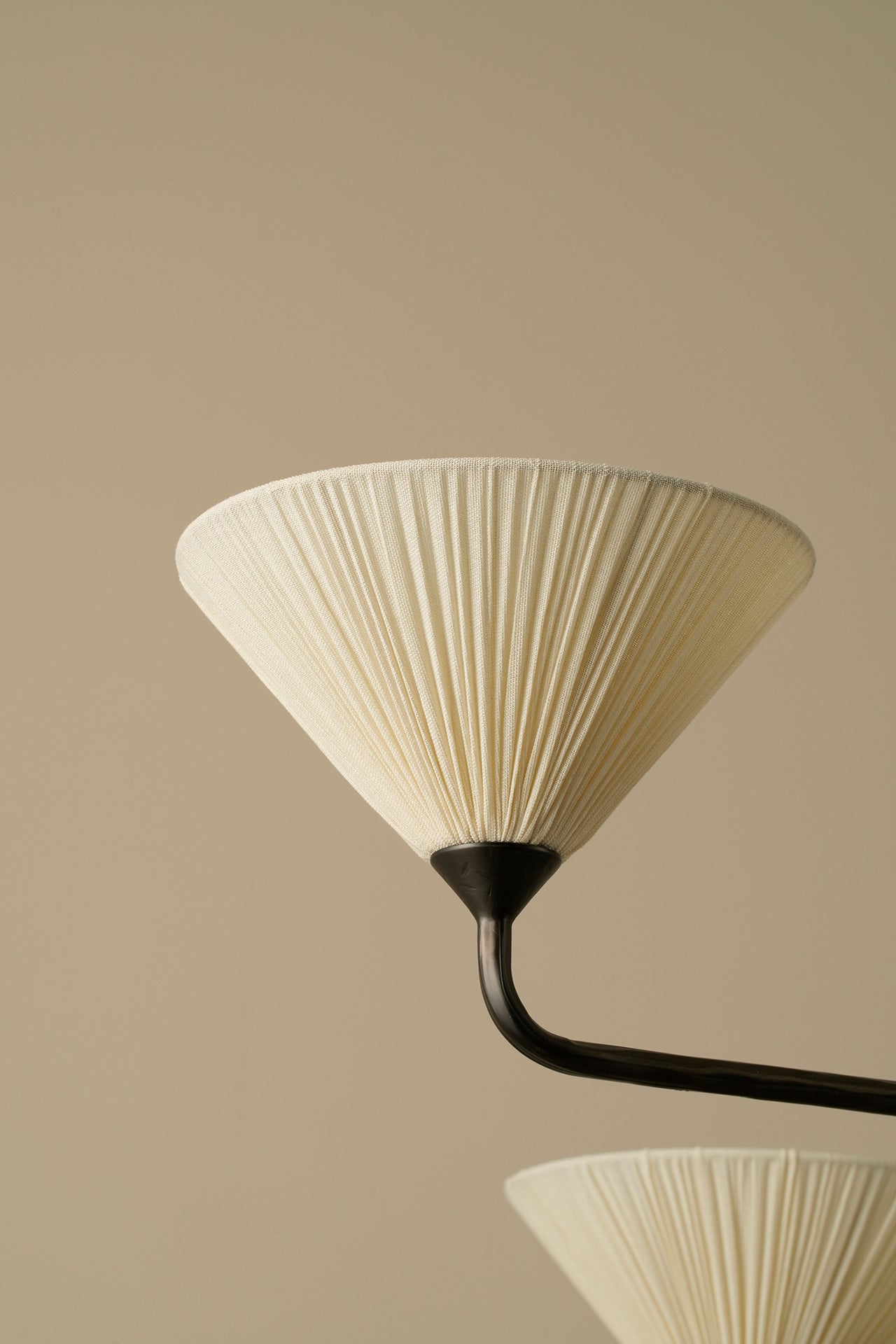 A close-up of a pleated cream-colored conical lampshade attached to a dark curved metal arm, set against a solid warm beige background.