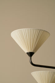 A close-up of a pleated cream-colored conical lampshade attached to a dark curved metal arm, set against a solid warm beige background.