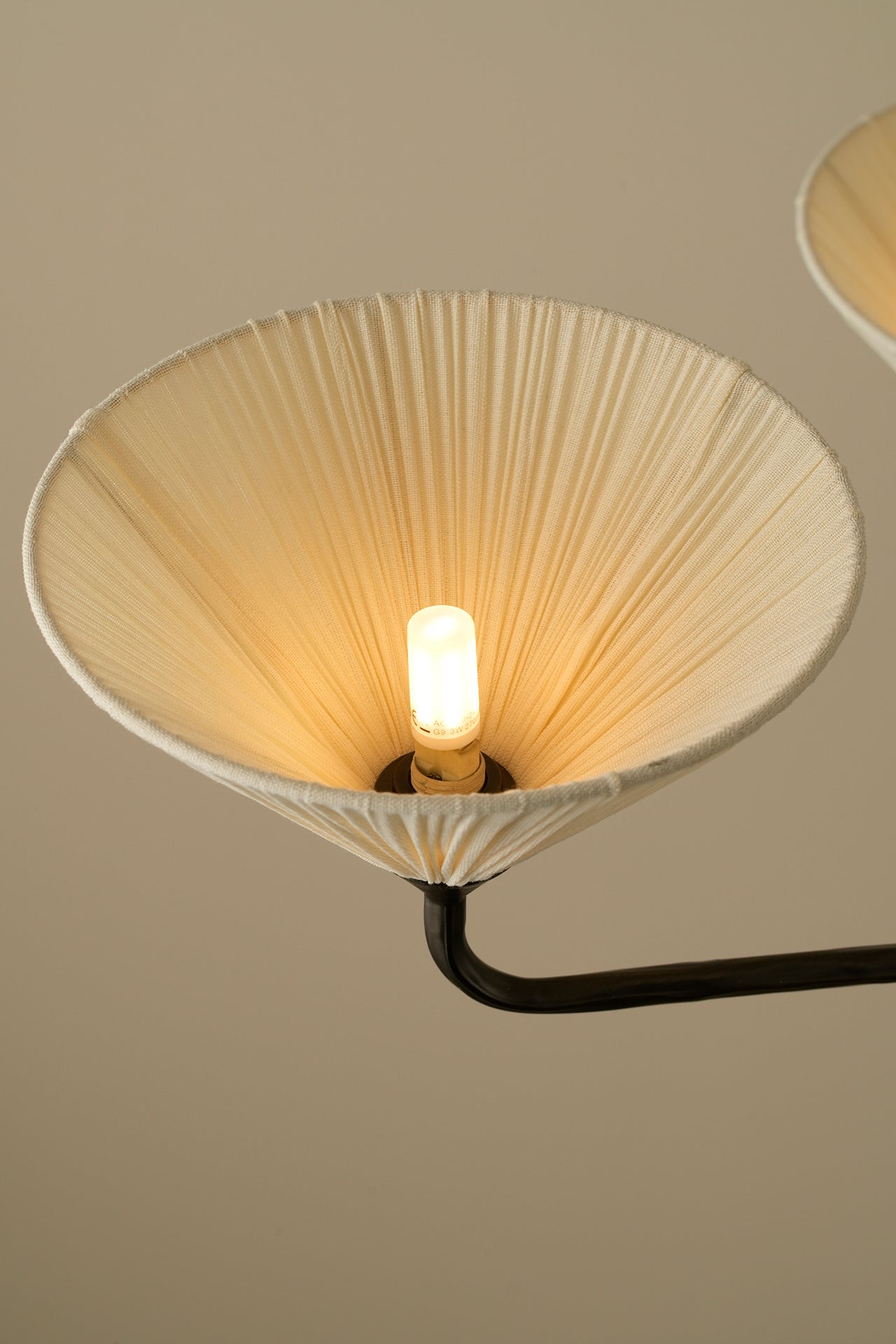 A close-up of a modern wall sconce featuring a large, conical pleated shade in a warm cream color, illuminated by a single central bulb, with a black metal arm extending against a solid beige background.