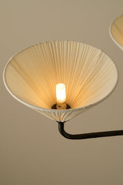 A close-up of a modern wall sconce featuring a large, conical pleated shade in a warm cream color, illuminated by a single central bulb, with a black metal arm extending against a solid beige background.