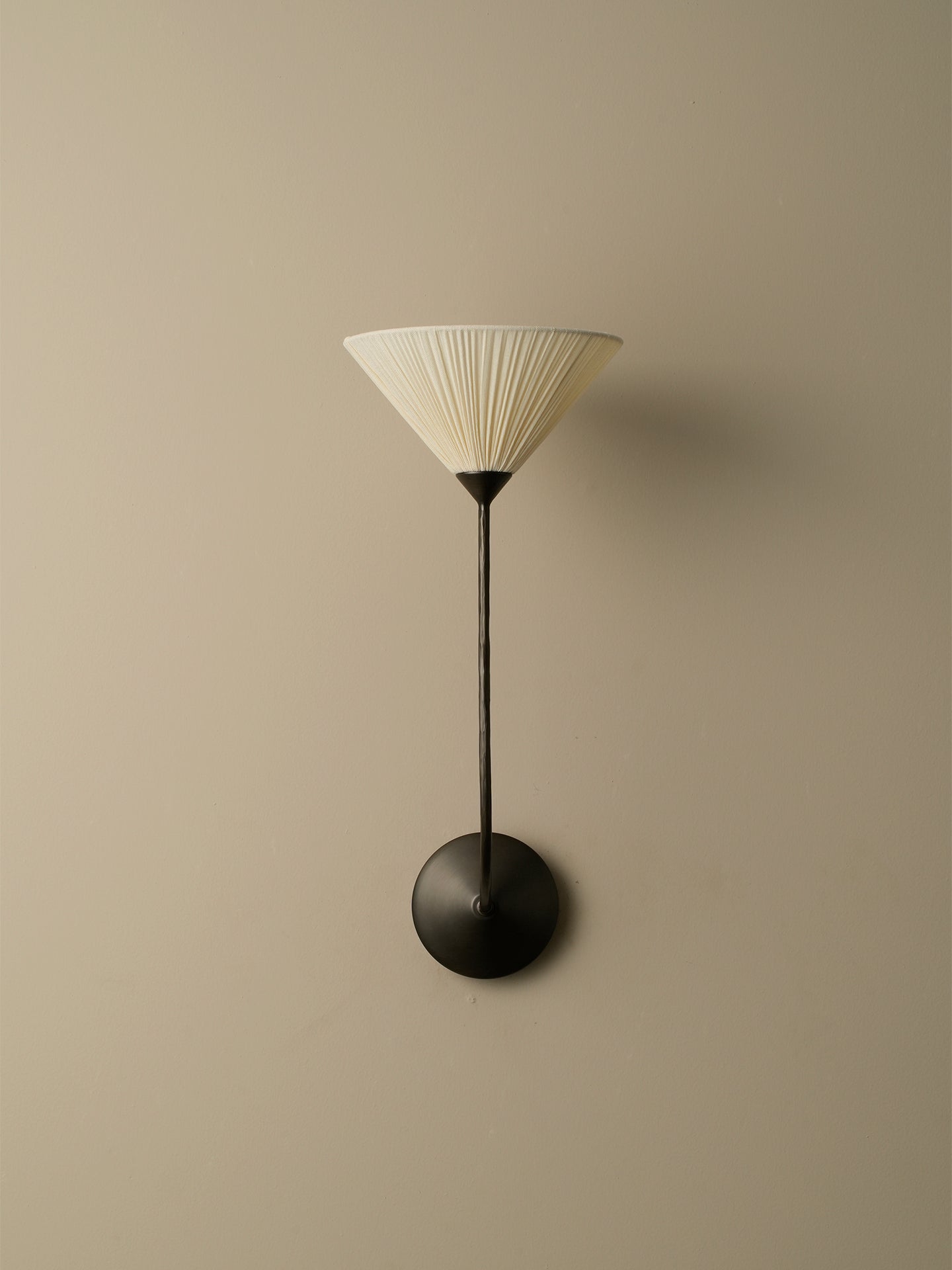 A modern wall-mounted sconce with a pleated off-white conical shade, a slim black stem, and a round black wall mount, shown against a plain beige wall.