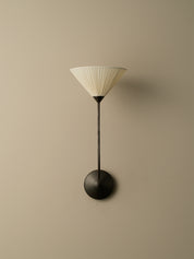 A modern wall-mounted sconce with a pleated off-white conical shade, a slim black stem, and a round black wall mount, shown against a plain beige wall.