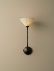 A modern wall sconce with a pleated white shade, a slim black stem, and a round black wall mount, shown against a plain beige wall.