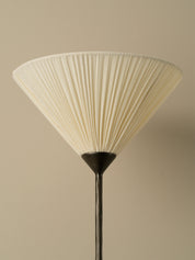 A close-up of a pleated fabric lampshade in a warm cream or off-white color, attached to a dark metal stand against a solid beige background.