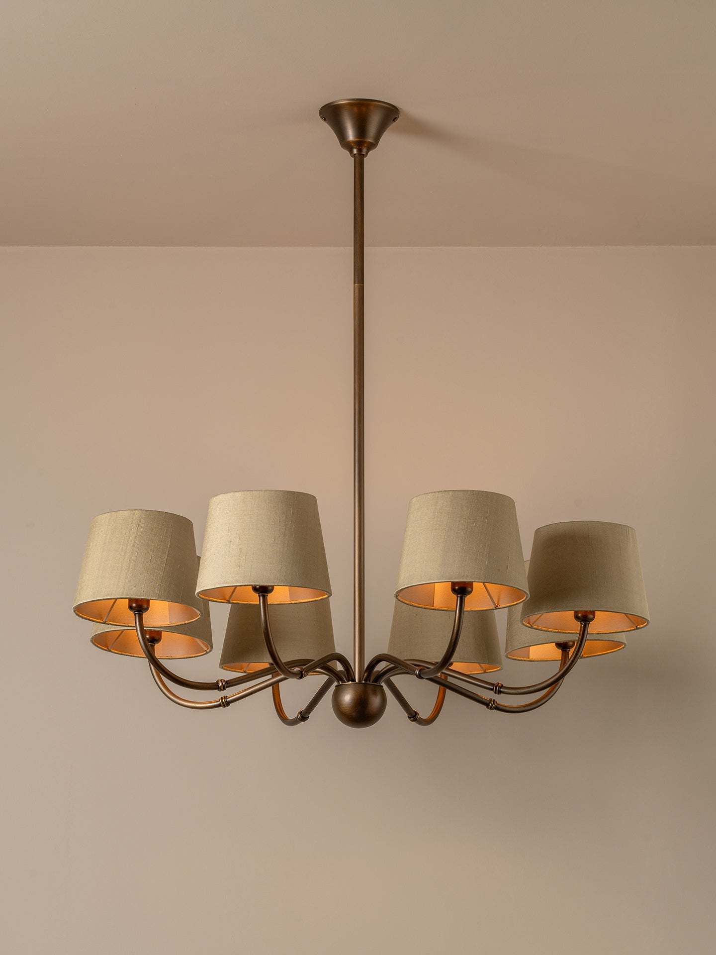 A six-light chandelier with a dark bronze metal frame and beige fabric drum shades, shown hanging against a neutral beige wall and ceiling.