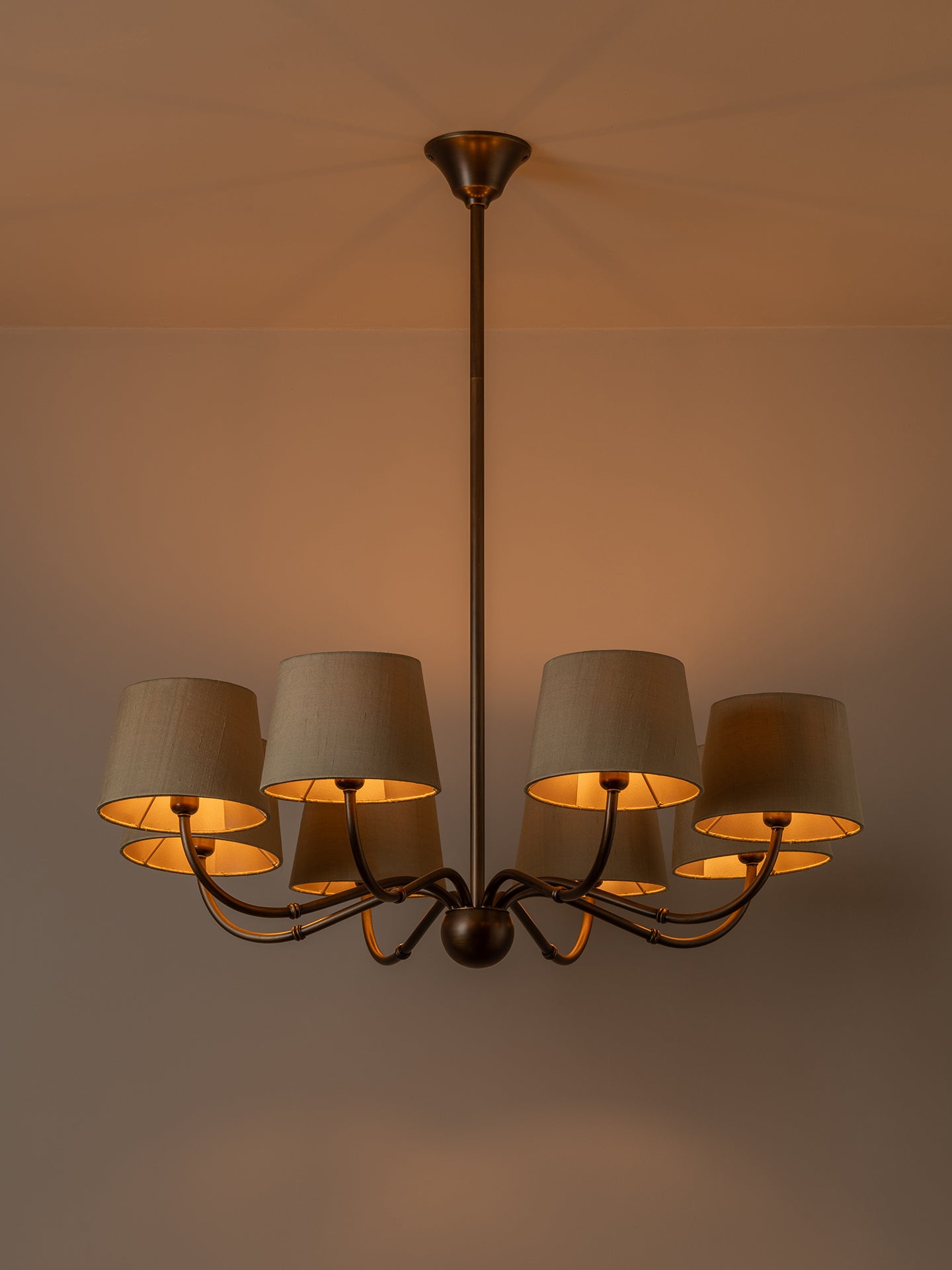 A multi-light chandelier with a dark bronze metal frame and six tapered beige fabric shades, shown illuminated against a warm beige wall and ceiling.