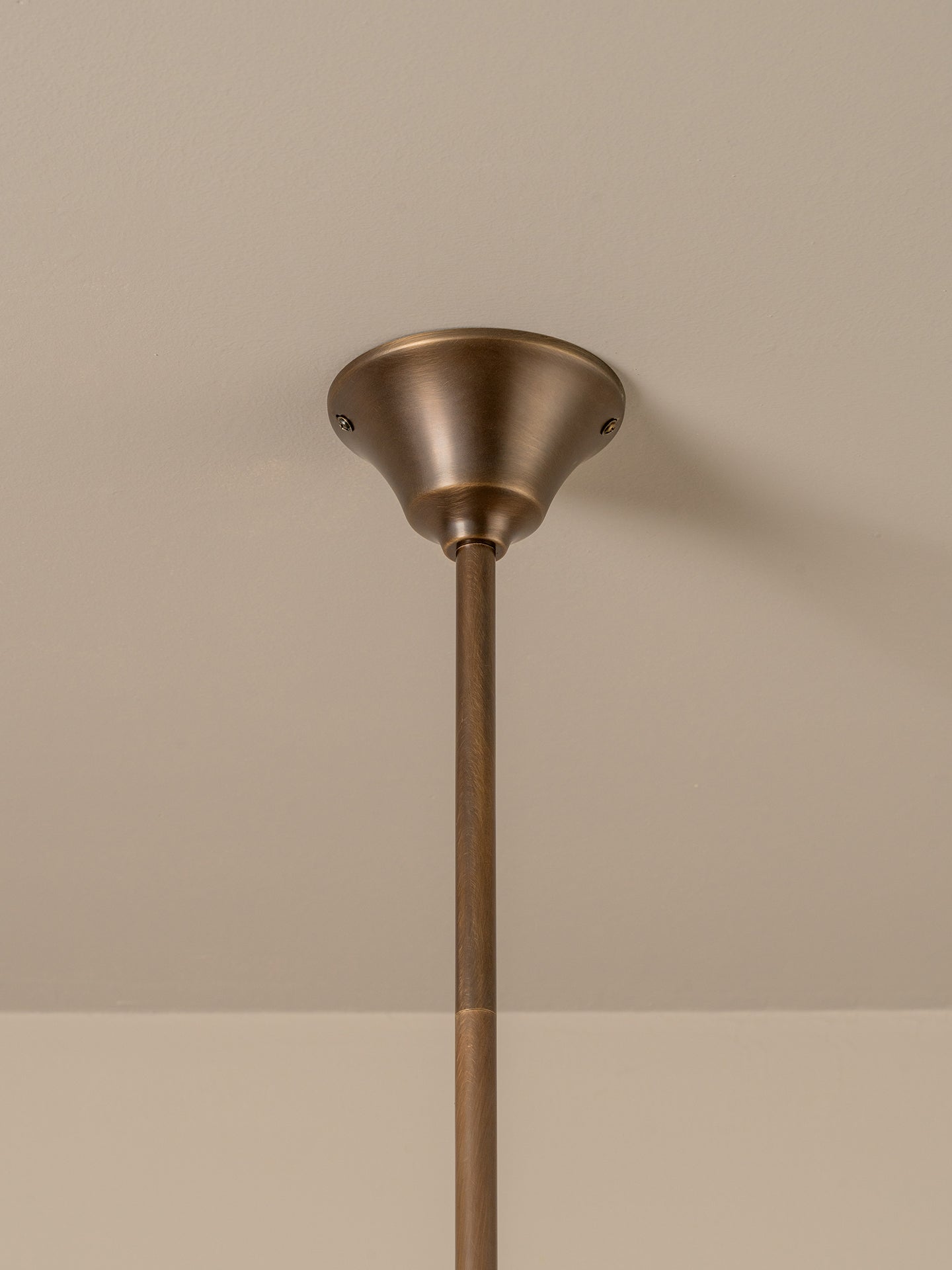 A close-up of a ceiling-mounted light fixture with a warm bronze-finished ceiling canopy and a matching vertical downrod against a neutral beige wall.