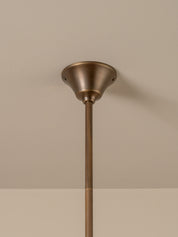A close-up of a ceiling-mounted light fixture with a warm bronze-finished ceiling canopy and a matching vertical downrod against a neutral beige wall.