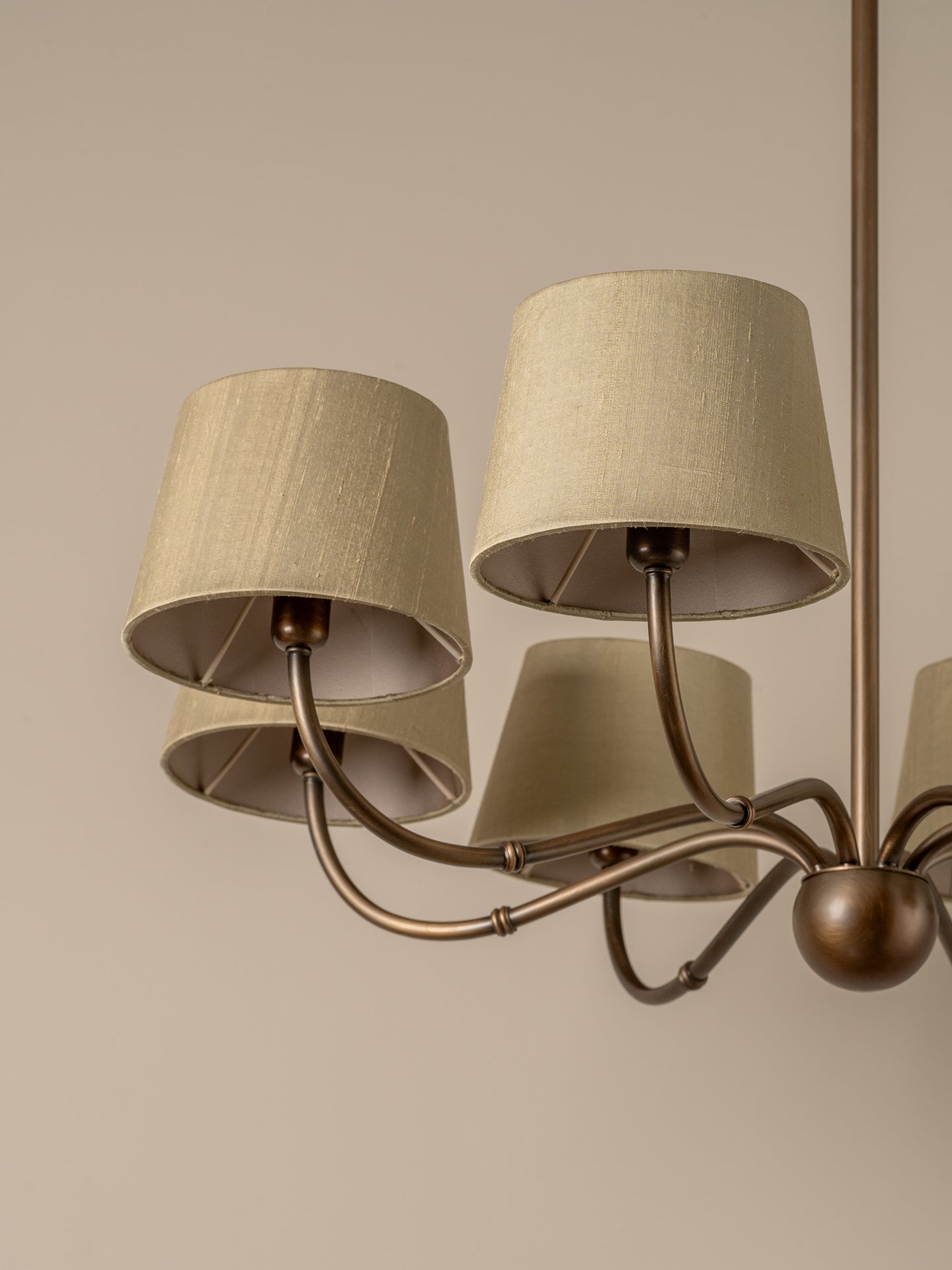 A close-up of a multi-arm chandelier with beige linen drum shades and a bronze-finished metal frame against a warm beige background.
