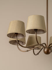 A close-up of a multi-arm chandelier with beige linen drum shades and a bronze-finished metal frame against a warm beige background.