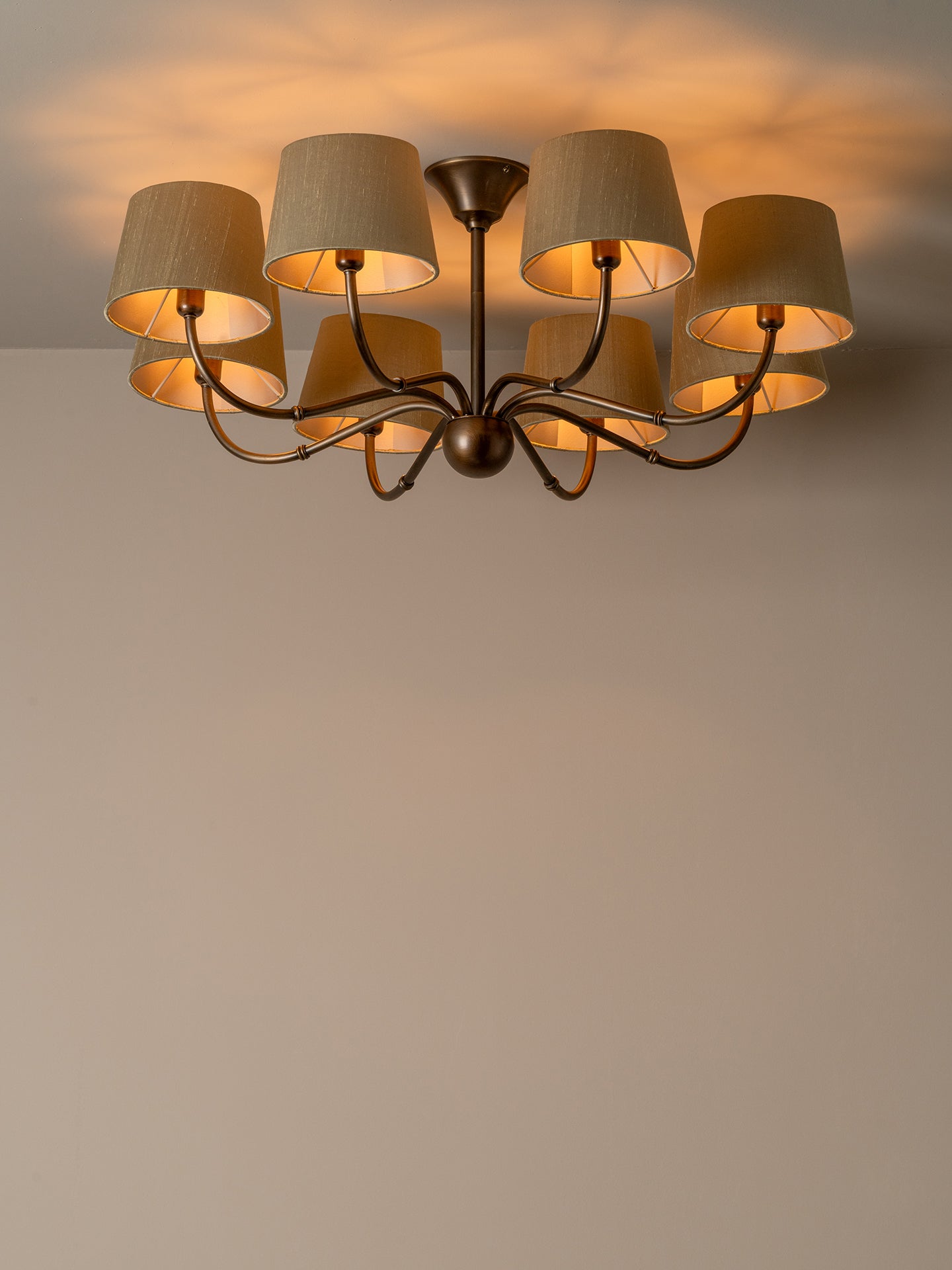 A multi-light ceiling fixture with six beige fabric drum shades and a dark bronze metal frame, shown illuminated against a plain beige wall.