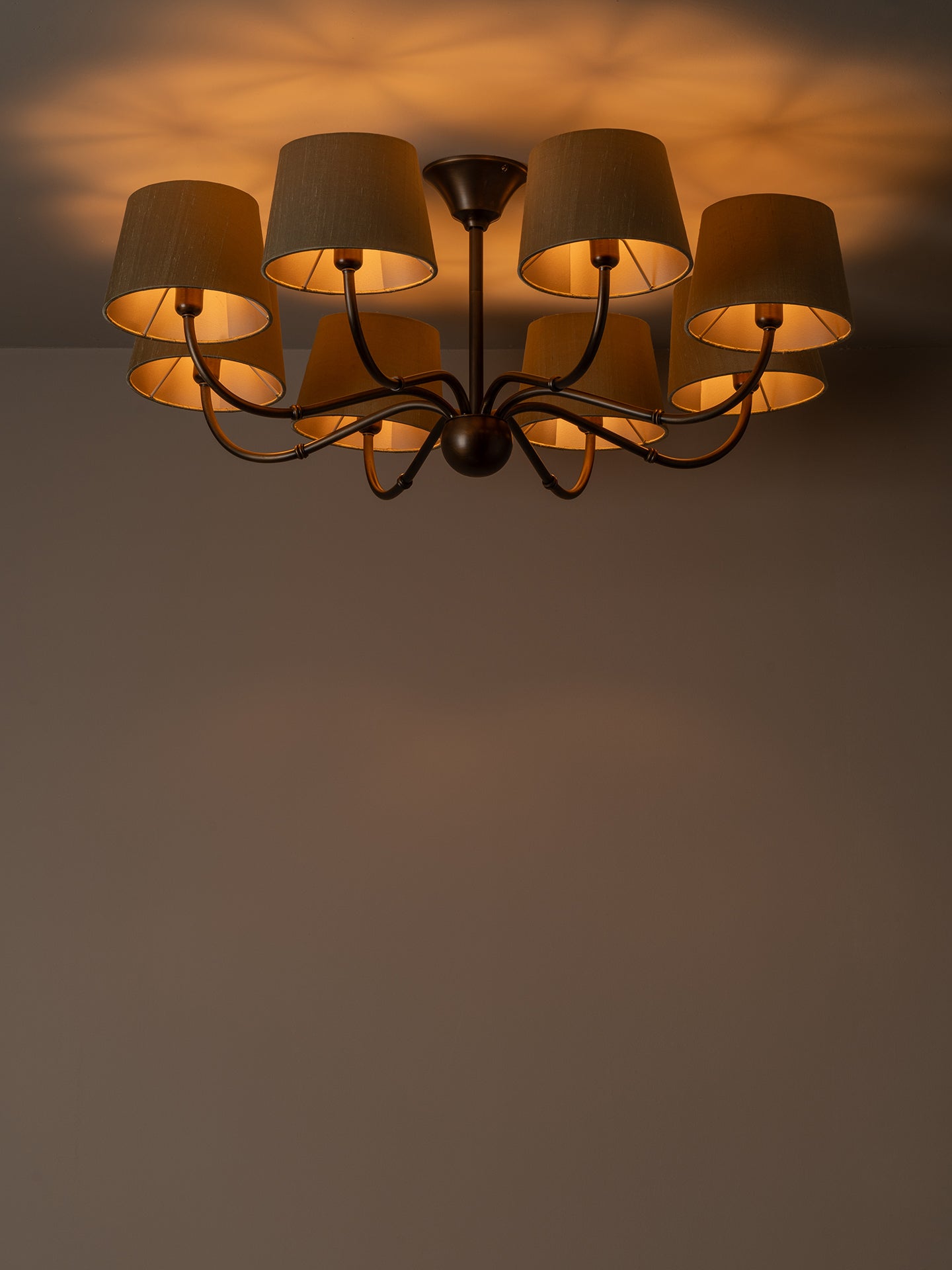 A multi-light chandelier with dark metal arms and beige fabric drum shades, shown illuminated against a warm brown wall and ceiling.
