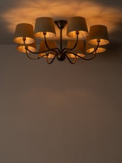 A multi-light chandelier with dark metal arms and beige fabric drum shades, shown illuminated against a warm brown wall and ceiling.
