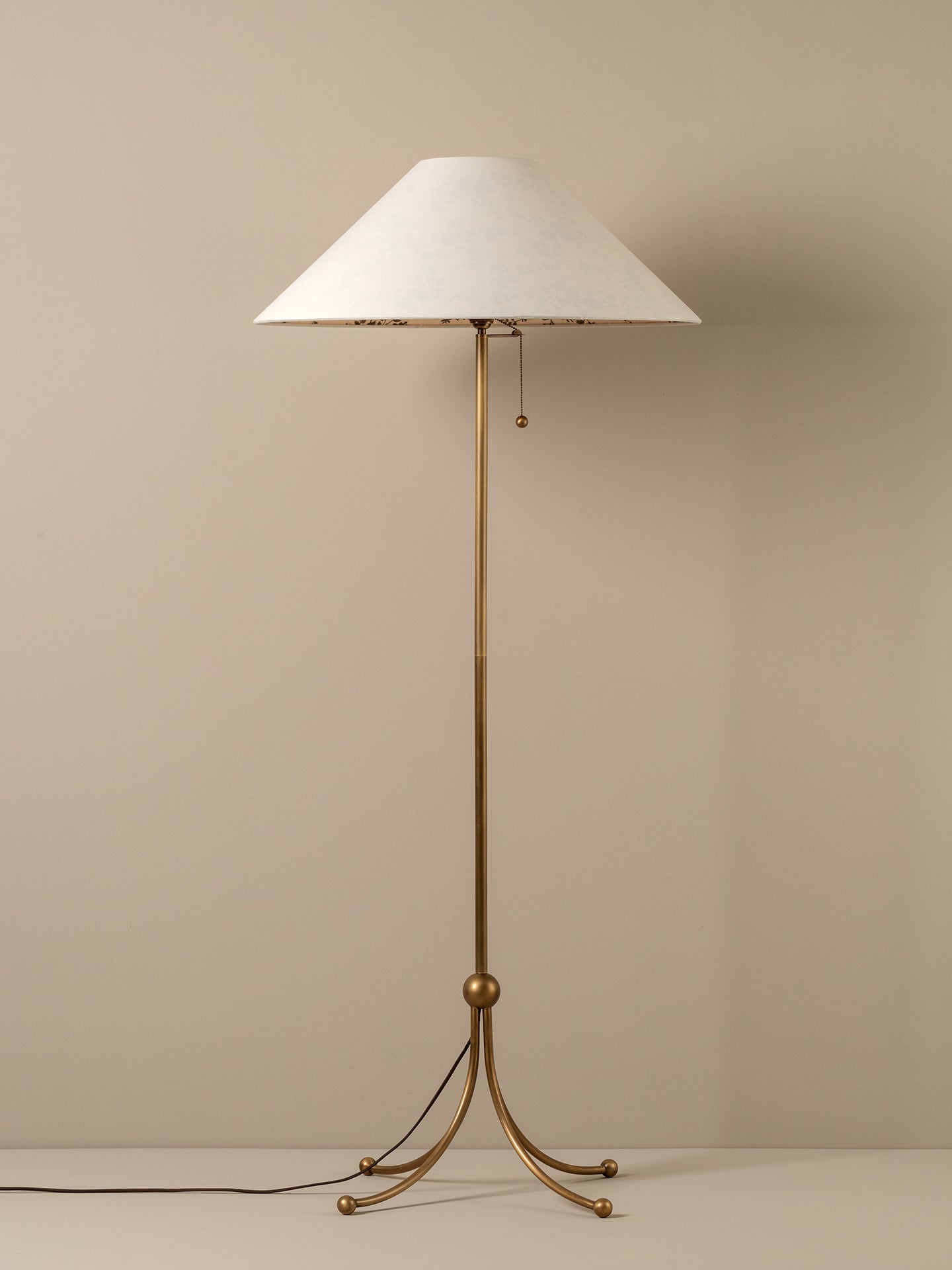 A mid-century modern floor lamp with a tapered white fabric shade and a polished brass tripod base, set against a neutral beige background.