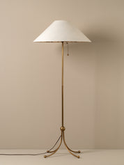 A mid-century modern floor lamp with a tapered white fabric shade and a polished brass tripod base, set against a neutral beige background.