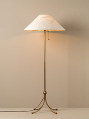 A floor lamp with a tapered off-white shade, a warm glowing light, and a brass-finished metal stand with three legs, set against a neutral beige background.