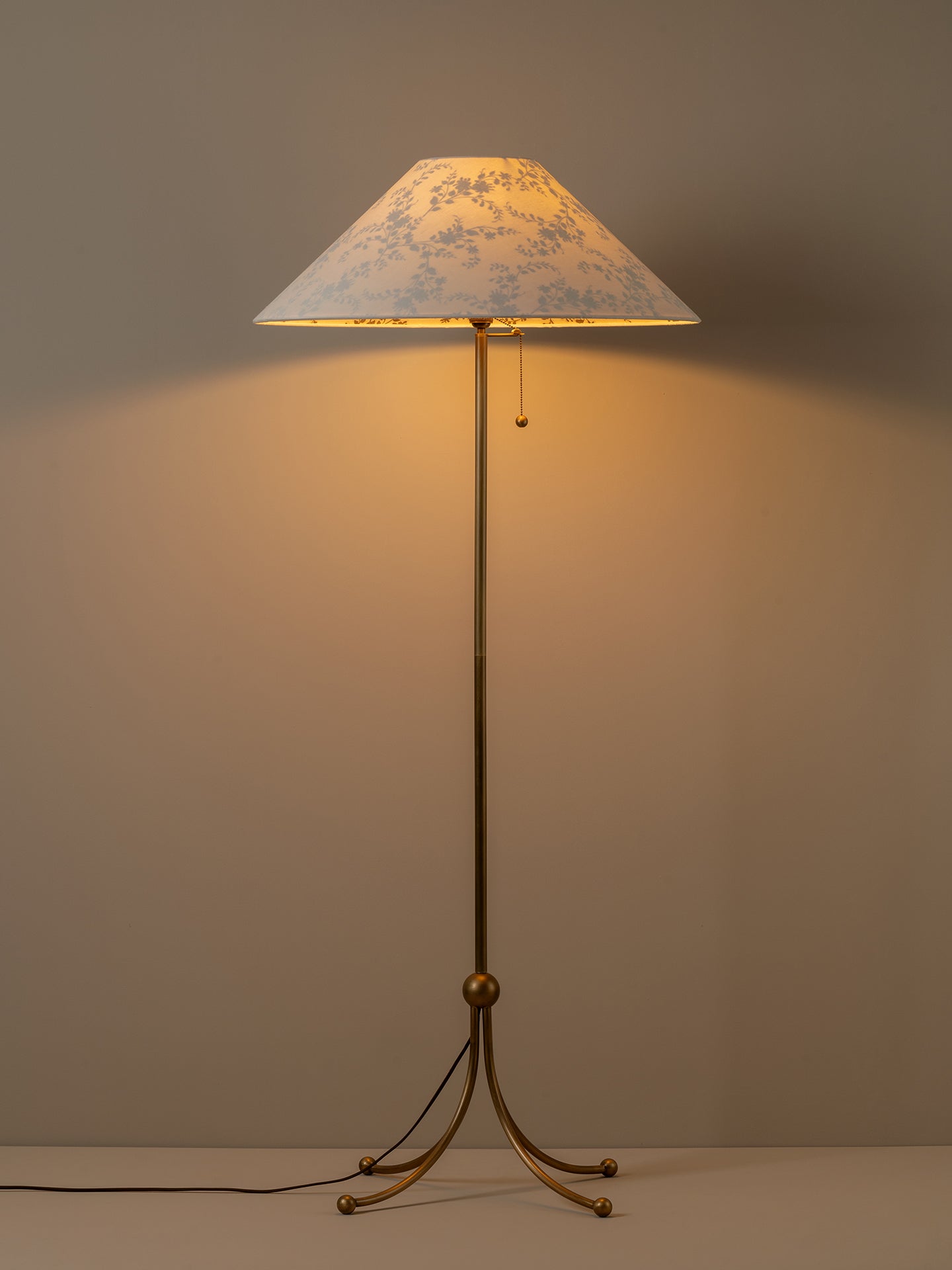 A vintage-style floor lamp with a warm brass-colored metal stand and a conical shade featuring a delicate blue floral pattern on a light background, illuminated against a neutral gray wall.