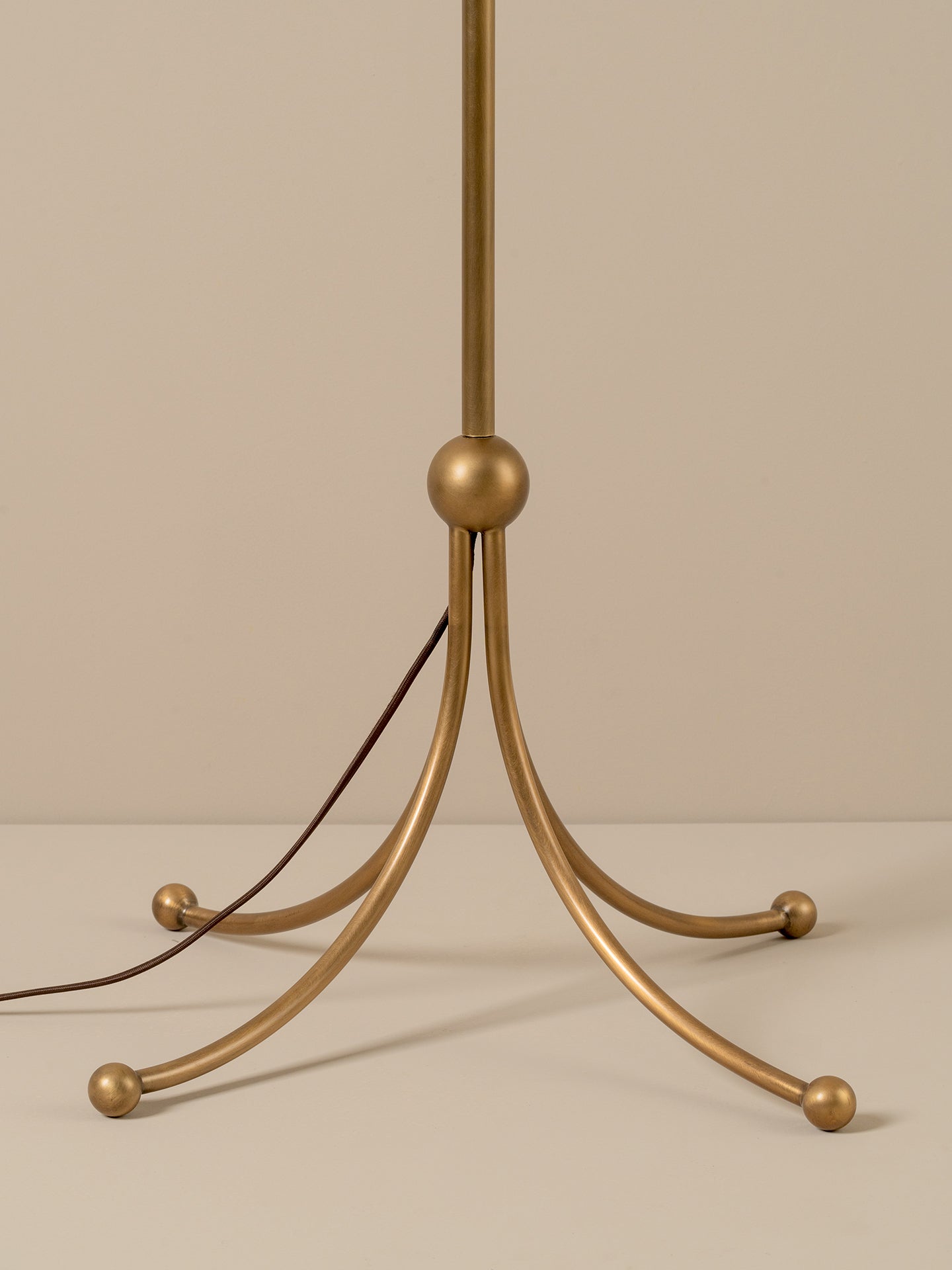 A close-up of a floor lamp base with a brushed brass/gold finish and a dark brown power cord, set against a solid beige background.