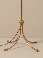 A close-up of a floor lamp base with a brushed brass/gold finish and a dark brown power cord, set against a solid beige background.