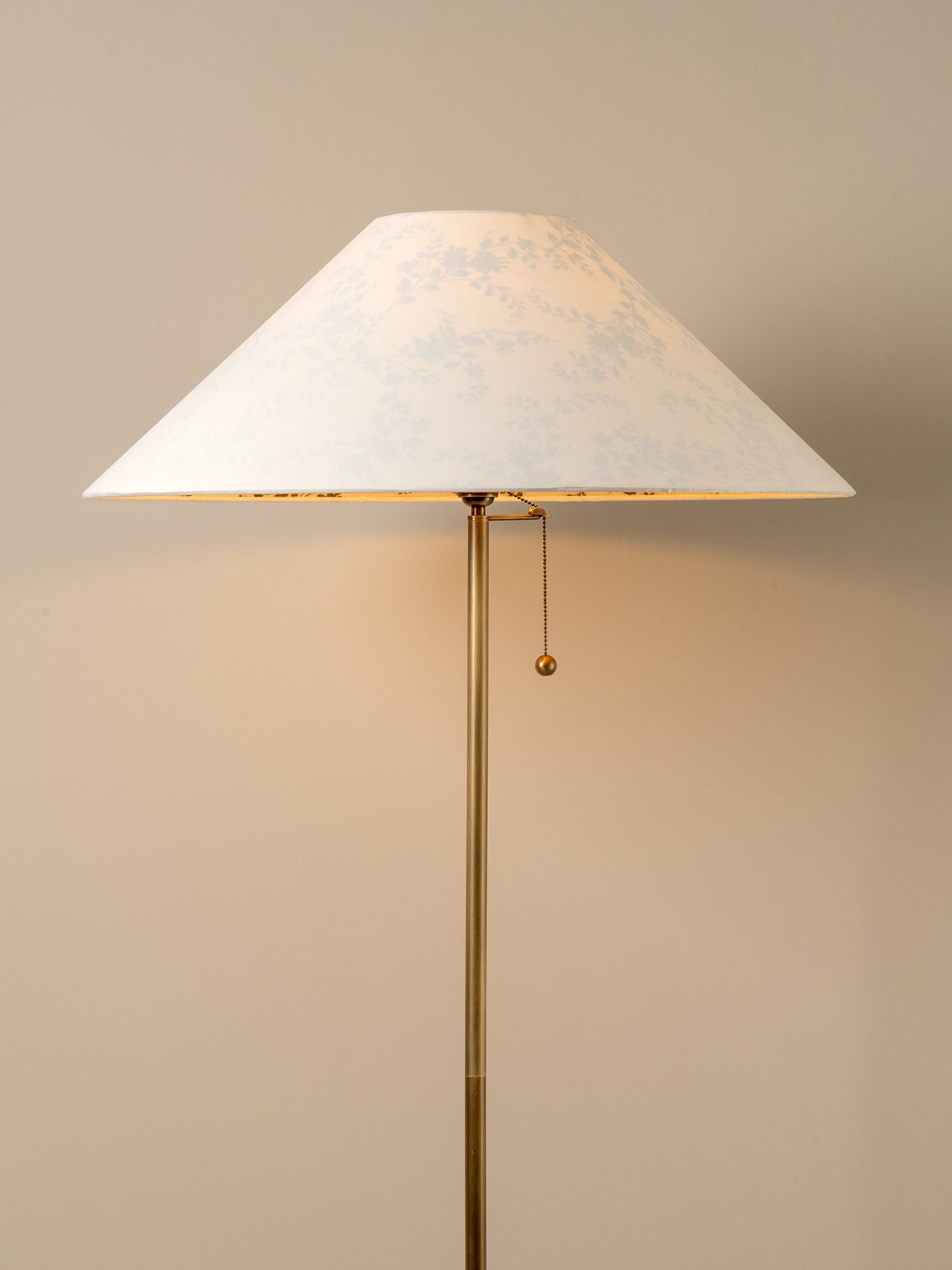 A floor lamp with a tapered white fabric shade featuring a subtle light blue floral pattern, supported by a slim brass pole with a pull chain.