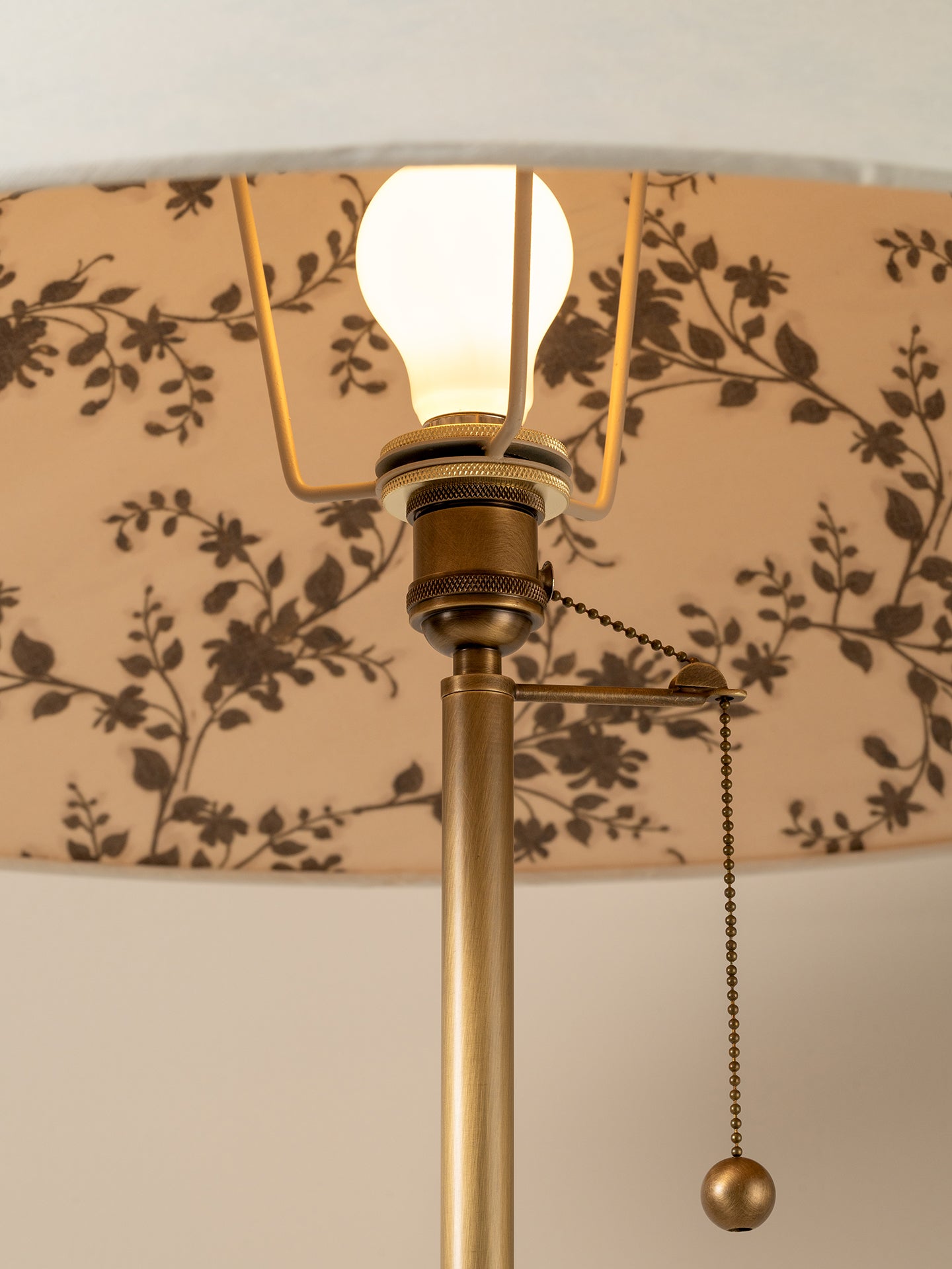 Close-up of a brass table lamp with a beige drum shade featuring a dark brown floral pattern, a glowing bulb, and a pull chain.