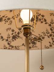 Close-up of a brass table lamp with a beige drum shade featuring a dark brown floral pattern, a glowing bulb, and a pull chain.