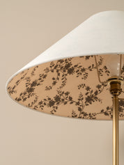 Close-up of a floor lamp shade with a white outer fabric and a beige inner panel featuring a dark brown floral vine pattern, mounted on a brass pole against a warm beige background.