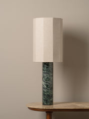 A table lamp with a textured beige fabric drum shade and a cylindrical dark green marble base, displayed on a wooden table against a neutral beige wall.