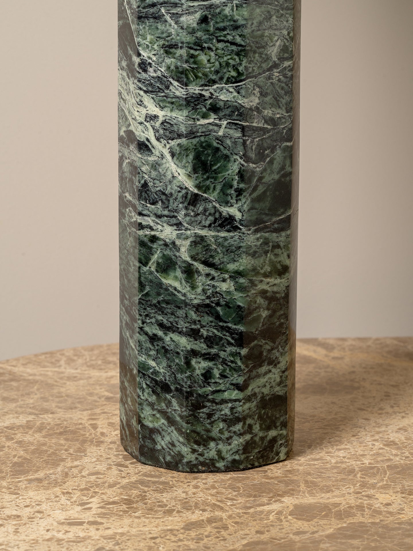 A cylindrical table lamp base made of polished green marble with natural white and dark veining, standing on a textured beige stone surface against a neutral beige background.