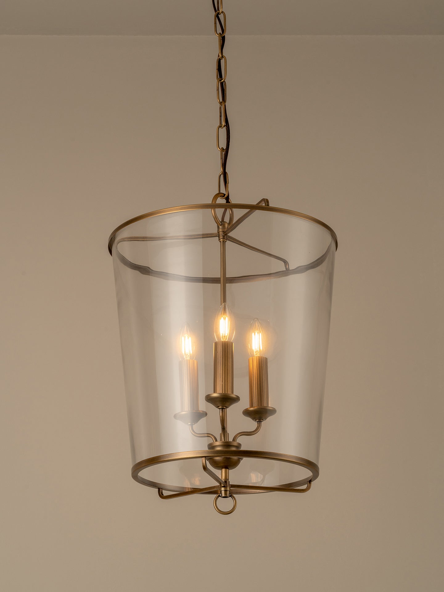 A hanging pendant light with a clear glass cylinder shade and an antique brass or bronze metal frame, suspended by a chain against a plain beige wall.