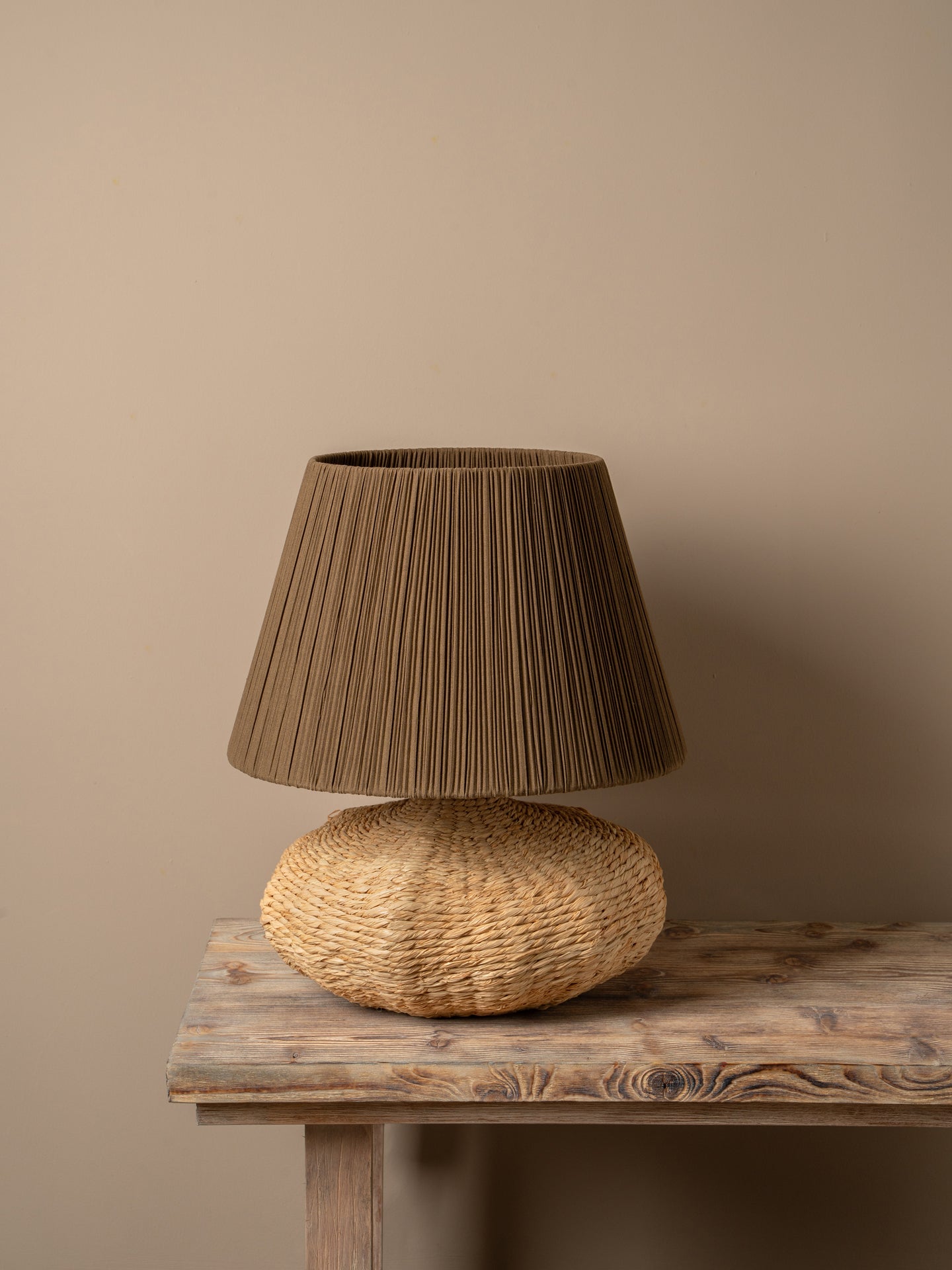A table lamp with a dark brown pleated fabric shade and a textured natural woven base, placed on a rustic wooden table against a warm neutral wall.